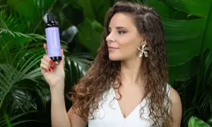 Une femme aux cheveux longs et bouclés tient un flacon de produit pour le soin des cheveux, entourée de verdure luxuriante.