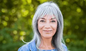 Une femme souriante aux cheveux gris argentés, un balayage subtil pour cacher les cheveux blancs, porte une veste en jean.
