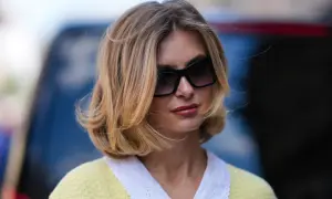 Une femme avec une coupe cheveux mi long blonde, des lunettes de soleil oversize et un pull jaune à col blanc.