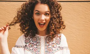 Une jeune femme surprise, sa coiffure cheveux bouclés tombant en cascade. Elle porte une blouse blanche brodée.