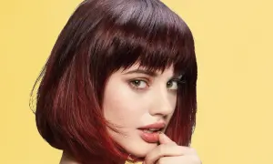Une jeune femme au regard intense arbore une coupe cheveux epais, mi-longue, dégradée et colorée de reflets auburn sur fond jaune vif.