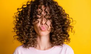 Une femme avec une magnifique coupe cheveux ondulés femme envoie un bisou, ses boucles brunes encadrant son visage sur fond jaune vif.