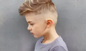 Un jeune garçon de 8 ans arbore une coupe de cheveux moderne avec un dégradé sur les côtés et des mèches blondes texturées sur le dessus.