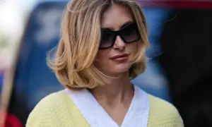 Une femme avec une coiffure sur le côté, cheveux mi-longs, porte des lunettes de soleil et un pull jaune à col blanc brodé.