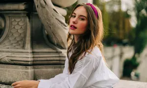 Jeune femme avec une coiffure avec serre tête fuchsia, chemise blanche, assise près d'une sculpture.
