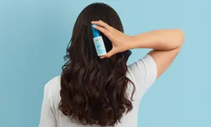 Une main tient un flacon de soin capillaire sur des cheveux bruns ondulés, pour lutter contre les cheveux déshydratés.