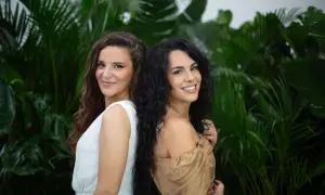 Deux femmes souriantes se tiennent dos à dos devant un feuillage luxuriant. L'une a des cheveux châtains ondulés, l'autre des cheveux noirs et bouclés, une magnifique chevelure mousseuse.