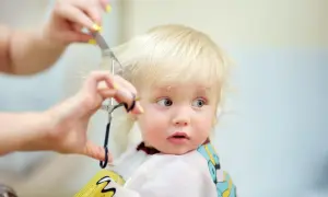 Un adorable petit garçon de 1 an, aux cheveux blonds, reçoit sa première coiffure. Ses grands yeux bleus regardent avec curiosité les ciseaux et le peigne.
