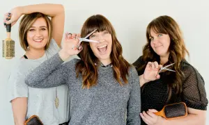 Trois femmes souriantes tiennent des ciseaux et des brosses. L'une d'elles joue avec ses ciseaux, comme pour savoir quand couper ses cheveux pour qu'ils poussent plus vite.