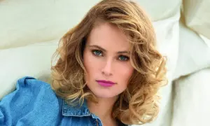 Une femme au visage rond arbore une coupe de cheveux mi-longs ondulés, mettant en valeur ses yeux bleus perçants.