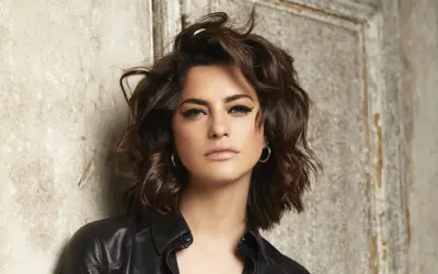 Une femme au regard intense, avec une coupe de cheveux pour visage carré qui encadre joliment ses traits. Elle porte une veste en cuir noire.