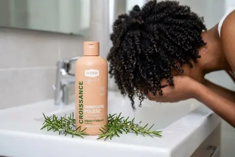 Shampoing "Croissance Pousse" WAAM, avec romarin, pour aider à comment faire pousser les cheveux. Une personne aux cheveux bouclés se lave le visage.