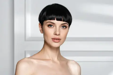 Une femme au regard intense arbore une coupe au bol audacieuse, ses cheveux noirs encadrant son visage aux traits fins.
