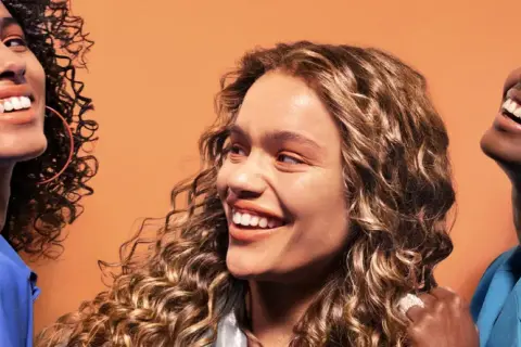 Trois femmes souriantes aux cheveux bouclés, chacune avec un style unique. Une inspiration pour savoir quelle frange pour quel visage.