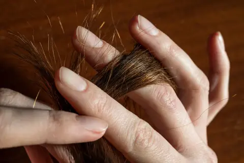 Des doigts effleurent une mèche de cheveux châtains, comme une **fourche à cheveux** naturelle, révélant des reflets dorés sous la lumière.