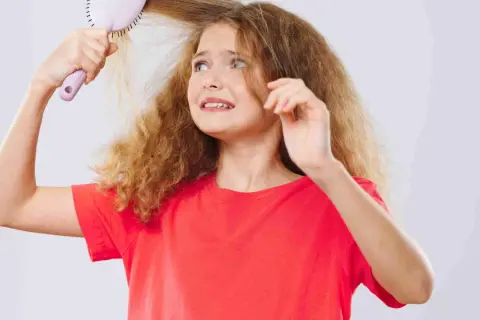Une jeune fille aux cheveux mousseux et fins essaie de se brosser les cheveux, l'air désemparée.