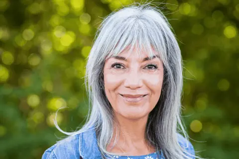 Une femme souriante aux cheveux gris argentés, un balayage subtil pour cacher les cheveux blancs, porte une veste en jean.