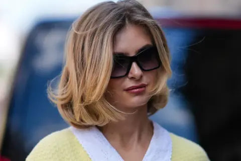 Une femme avec une coupe cheveux mi long blonde, des lunettes de soleil oversize et un pull jaune à col blanc.