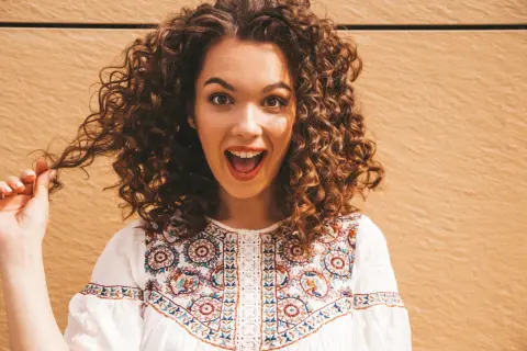 Une jeune femme surprise, sa coiffure cheveux bouclés tombant en cascade. Elle porte une blouse blanche brodée.