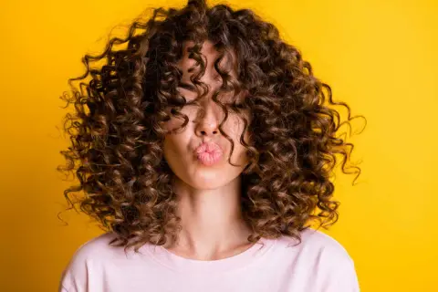 Une femme avec une magnifique coupe cheveux ondulés femme envoie un bisou, ses boucles brunes encadrant son visage sur fond jaune vif.
