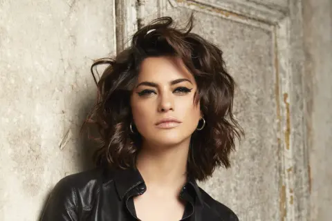 Une femme au regard intense, avec une coupe de cheveux pour visage carré qui encadre joliment ses traits. Elle porte une veste en cuir noire.