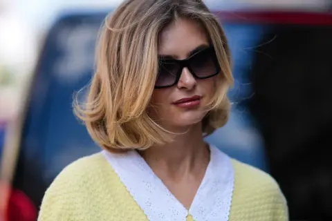 Une femme avec une coiffure sur le côté, cheveux mi-longs, porte des lunettes de soleil et un pull jaune à col blanc brodé.
