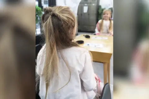 Une petite fille avec une jolie demi queue de cheval torsadée, assise à une table.