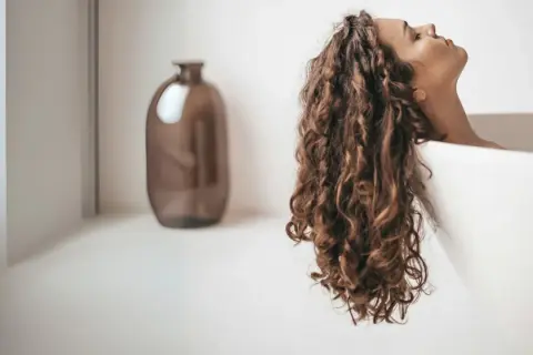 Une femme aux cheveux bouclés se détend dans une baignoire. Apprenez comment utiliser un après-shampoing pour des boucles douces et brillantes.