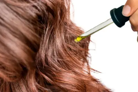 Une goutte d'huile dorée tombe d'une pipette sur des cheveux châtain clair, promettant soin et brillance.