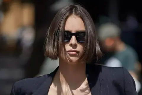 Une femme porte des lunettes de soleil et une veste, ses cheveux arborent une coupe carré court dégradé effilé.