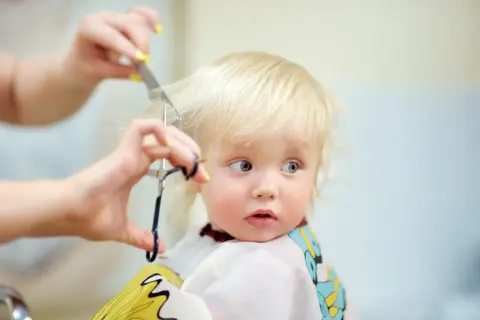 Un adorable petit garçon de 1 an, aux cheveux blonds, reçoit sa première coiffure. Ses grands yeux bleus regardent avec curiosité les ciseaux et le peigne.
