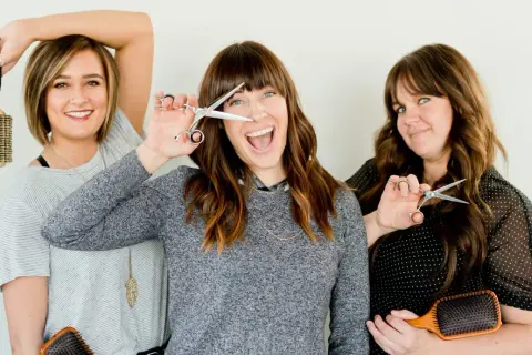 Trois femmes souriantes tiennent des ciseaux et des brosses. L'une d'elles joue avec ses ciseaux, comme pour savoir quand couper ses cheveux pour qu'ils poussent plus vite.