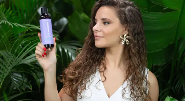 Une femme aux cheveux longs et bouclés tient un flacon de produit pour le soin des cheveux, entourée de verdure luxuriante.