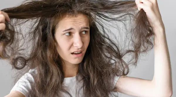 Jeune femme aux cheveux bruns et indisciplinés, se demandant comment faire pour que les cheveux ne gonflent pas. Elle tire sur une mèche, l'air frustrée.