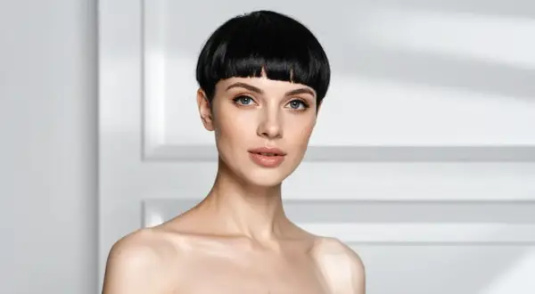 Une femme au regard intense arbore une coupe au bol audacieuse, ses cheveux noirs encadrant son visage aux traits fins.