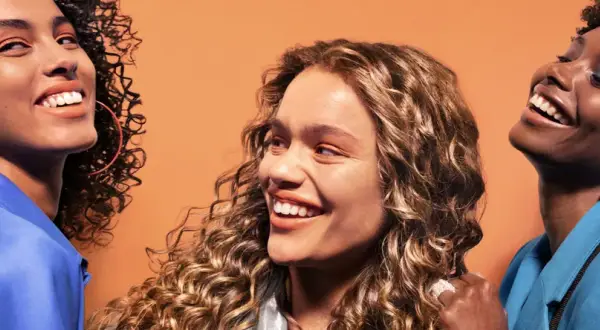 Trois femmes souriantes aux cheveux bouclés, chacune avec un style unique. Une inspiration pour savoir quelle frange pour quel visage.