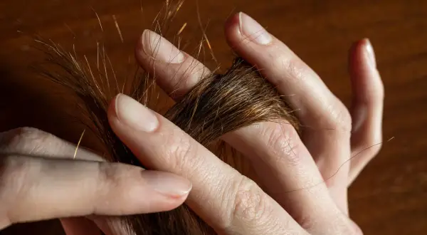 Des doigts effleurent une mèche de cheveux châtains, comme une **fourche à cheveux** naturelle, révélant des reflets dorés sous la lumière.