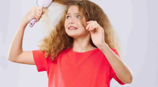 Une jeune fille aux cheveux mousseux et fins essaie de se brosser les cheveux, l'air désemparée.