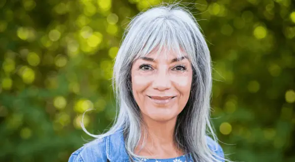 Une femme souriante aux cheveux gris argentés, un balayage subtil pour cacher les cheveux blancs, porte une veste en jean.