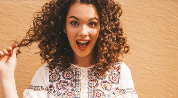 Une jeune femme surprise, sa coiffure cheveux bouclés tombant en cascade. Elle porte une blouse blanche brodée.