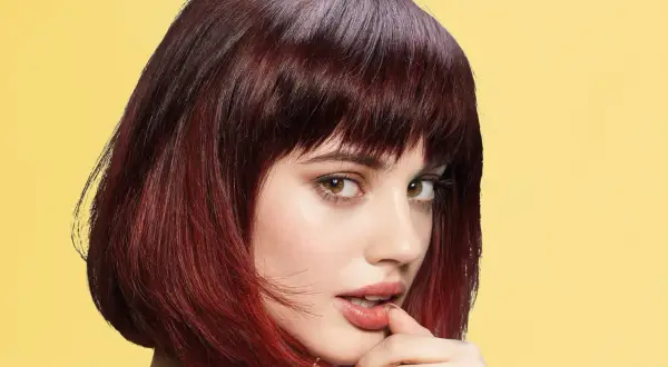 Une jeune femme au regard intense arbore une coupe cheveux epais, mi-longue, dégradée et colorée de reflets auburn sur fond jaune vif.