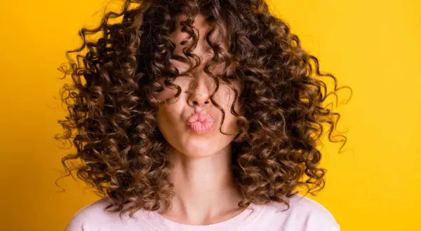 Une femme avec une magnifique coupe cheveux ondulés femme envoie un bisou, ses boucles brunes encadrant son visage sur fond jaune vif.