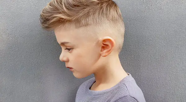 Un jeune garçon de 8 ans arbore une coupe de cheveux moderne avec un dégradé sur les côtés et des mèches blondes texturées sur le dessus.