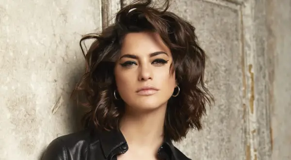 Une femme au regard intense, avec une coupe de cheveux pour visage carré qui encadre joliment ses traits. Elle porte une veste en cuir noire.