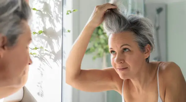 Une femme regarde ses cheveux poivre et sel dans le miroir, se demandant comment les blanchir pour un look plus lumineux.