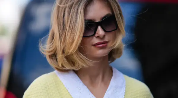Une femme avec une coiffure sur le côté, cheveux mi-longs, porte des lunettes de soleil et un pull jaune à col blanc brodé.