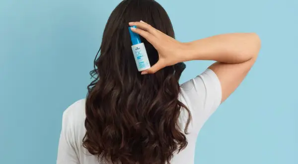 Une main tient un flacon de soin capillaire sur des cheveux bruns ondulés, pour lutter contre les cheveux déshydratés.