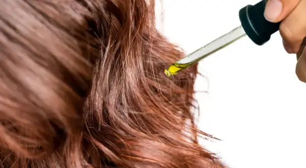 Une goutte d'huile dorée tombe d'une pipette sur des cheveux châtain clair, promettant soin et brillance.