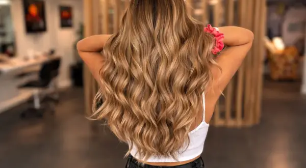 Chevelure longue et ondulée, aux reflets blonds subtils, parfaite pour éclaircir ses cheveux colorés. Un chouchou rose orne le poignet.