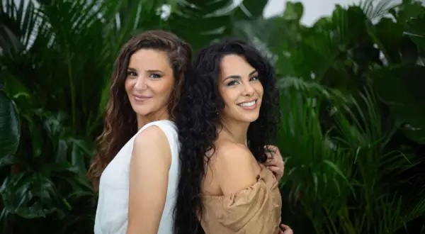 Deux femmes souriantes se tiennent dos à dos devant un feuillage luxuriant. L'une a des cheveux châtains ondulés, l'autre des cheveux noirs et bouclés, une magnifique chevelure mousseuse.